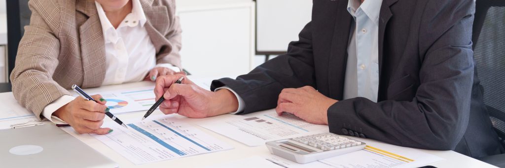 Two people reviewing documents at a desk with a calculator, a tablet, and a laptop.